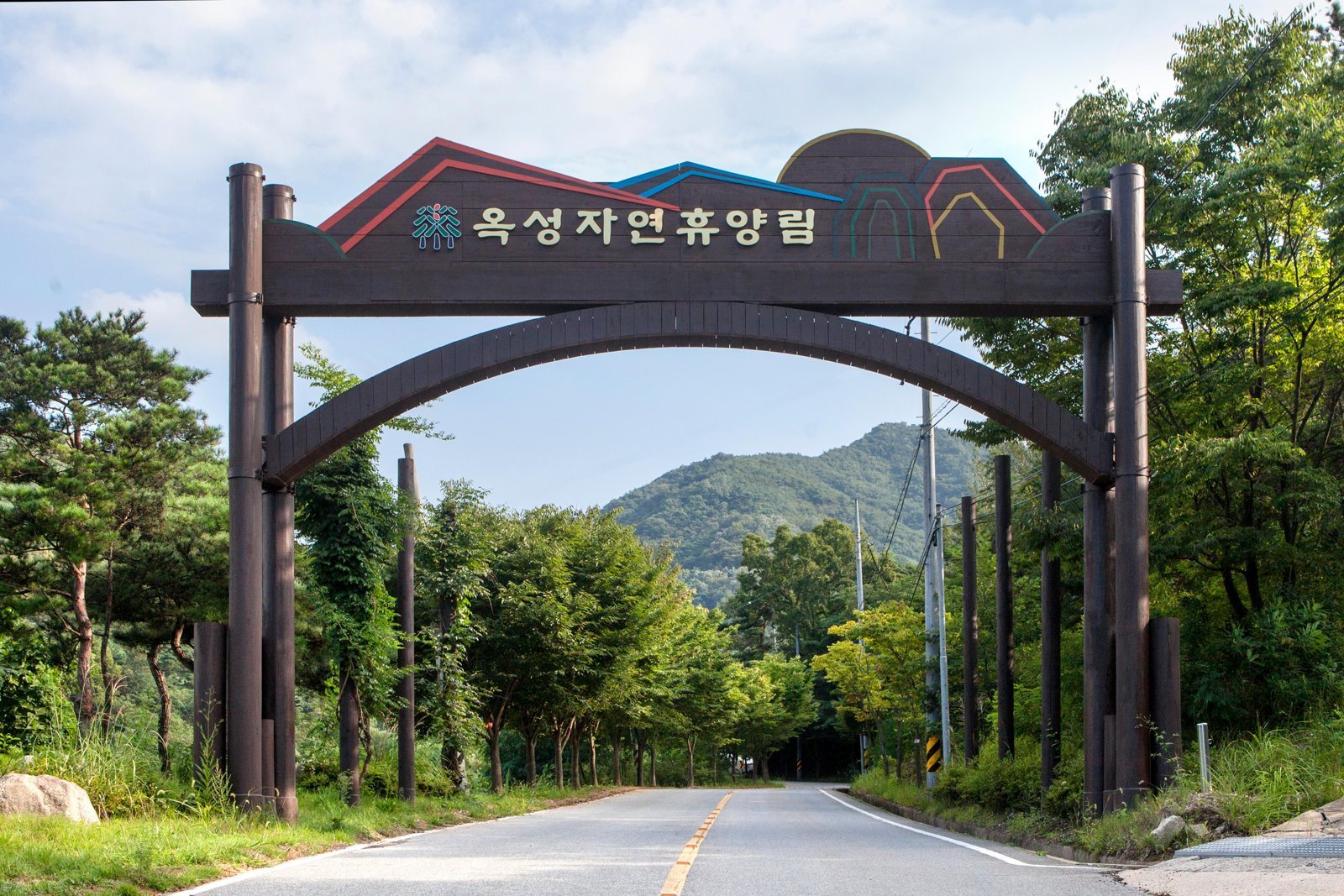 «옥성자연휴양림» 글자가 붙은 나무 입구 아치와 숲 사이로 난 길, 뒤로 초록 산
