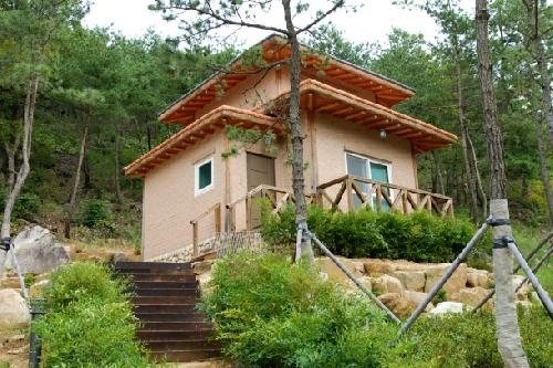 돌 축대 위 2층 숲속의 집(밝은 벽, 나무 난간 데크), 앞쪽 나무 계단과 소나무
